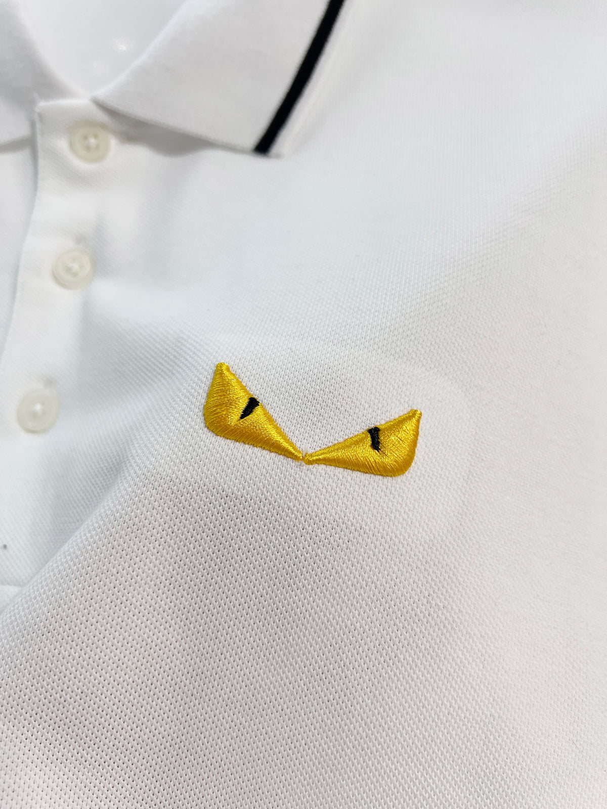 A polo shirt with embroidered letters on the eyes of a golden little monster on the chest